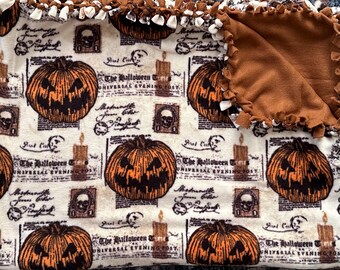 Può includere: Una coperta in pile a tema Halloween con un motivo ripetuto di zucche arancioni, candele e teschi su sfondo crema. La coperta ha un bordo marrone con bordi annodati. Il testo "The Halloween Universal Evening Post" è visibile.