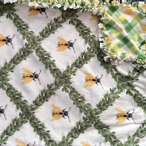 May include: A white fleece blanket with a repeating pattern of yellow and black bees and green leaf vines. The blanket has a green and yellow plaid corner and a knotted edge.