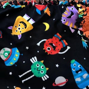 May include: A black fleece blanket featuring colorful cartoon space creatures, rockets, planets, and stars. The blanket has an orange border with knotted fringe. The design includes yellow, blue, red, green, and purple creatures.