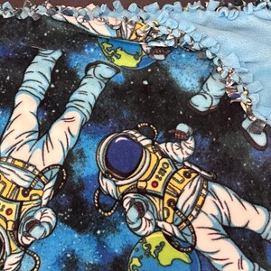 May include: A blue and black fleece blanket featuring a space theme. The blanket has a repeating pattern of astronauts, planets, and stars. The edges are trimmed with a light blue, knotted fringe.