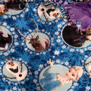 May include: A blue fleece blanket featuring characters from the animated movie Frozen. The blanket has a repeating pattern of snowflakes and circular images of Anna, Elsa, Olaf, and Sven. The edges are finished with a purple fringe.