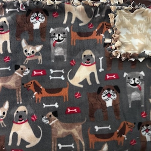 May include: A gray fleece blanket featuring a repeating pattern of various dog breeds in brown, tan, and gray. The blanket includes dog bones and red food bowls. A corner of the blanket is folded over, revealing a tan and white star pattern.