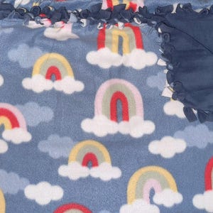 May include: A blue fleece blanket with a repeating pattern of colorful rainbows and white clouds. The blanket has a navy blue border with knotted fringe. The rainbows feature red, yellow, and pink arches.
