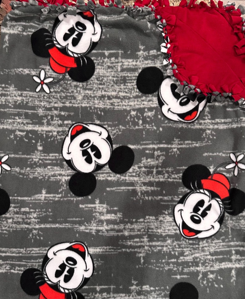 Puede incluir: Una manta de forro polar con fondo gris y un patr&oacute;n de rayas horizontales blancas desgastadas. La manta est&aacute; decorada con ilustraciones repetidas de Minnie Mouse con un sombrero rojo y una flor blanca, con un borde de flecos anudados.