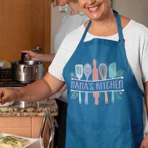 May include: A blue kitchen apron with a white background and the text "Nana's Kitchen" in a white banner. The apron has a pocket and features a design of colorful kitchen utensils with floral accents.