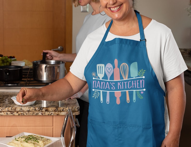 May include: A blue kitchen apron with a white background and the text "Nana's Kitchen" in a white banner. The apron has a pocket and features a design of colorful kitchen utensils with floral accents.