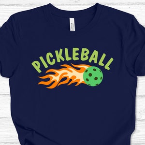 May include: A navy blue t-shirt featuring a green and orange graphic of a pickleball on fire with the text "Pickleball" in green.