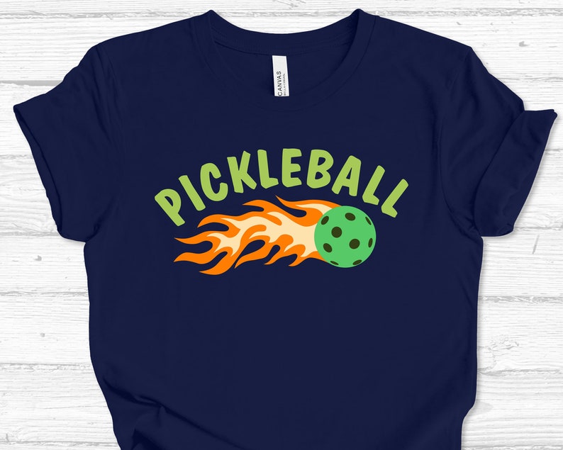 May include: A navy blue t-shirt featuring a green and orange graphic of a pickleball on fire with the text "Pickleball" in green.