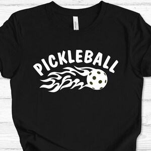 May include: Black t-shirt with a white graphic of a pickleball on fire with the text "Pickleball" above it.