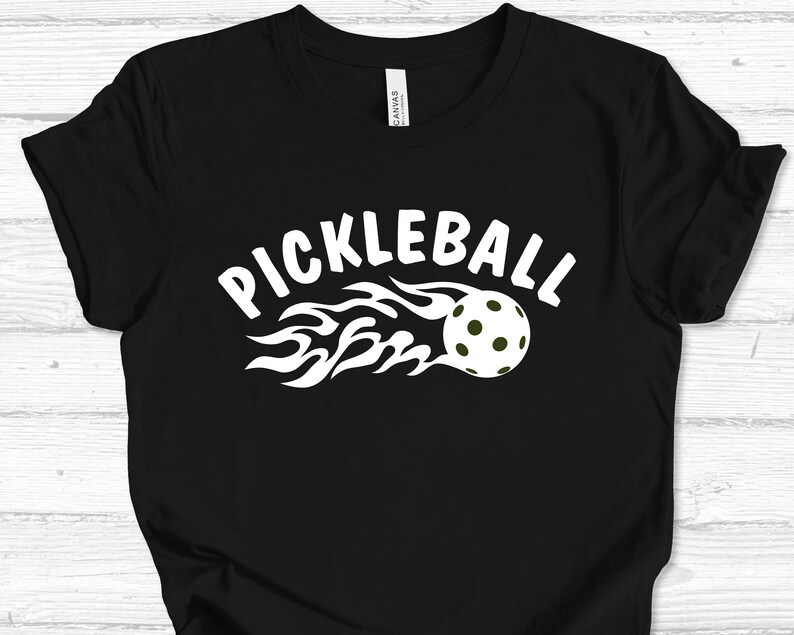 May include: Black t-shirt with a white graphic of a pickleball on fire with the text "Pickleball" above it.