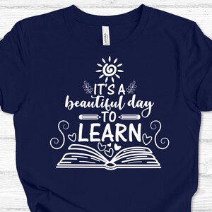 May include: A navy blue t-shirt with a white design featuring a sun, a book, pencils, hearts, and the text "It's a beautiful day to learn."