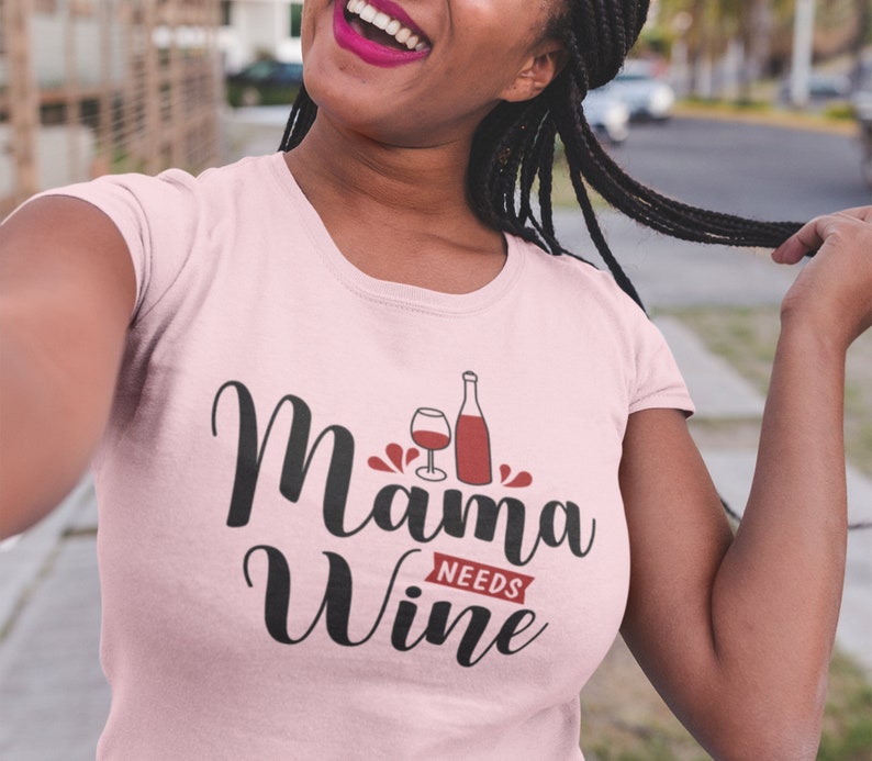 May include: Pink t-shirt with the text "Mama Needs Wine" in black cursive font. A glass of wine and a bottle of wine are depicted in red.