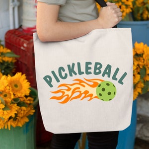 May include: A white canvas tote bag with a green and orange graphic of a pickleball on fire and the text "PICKLEBALL".