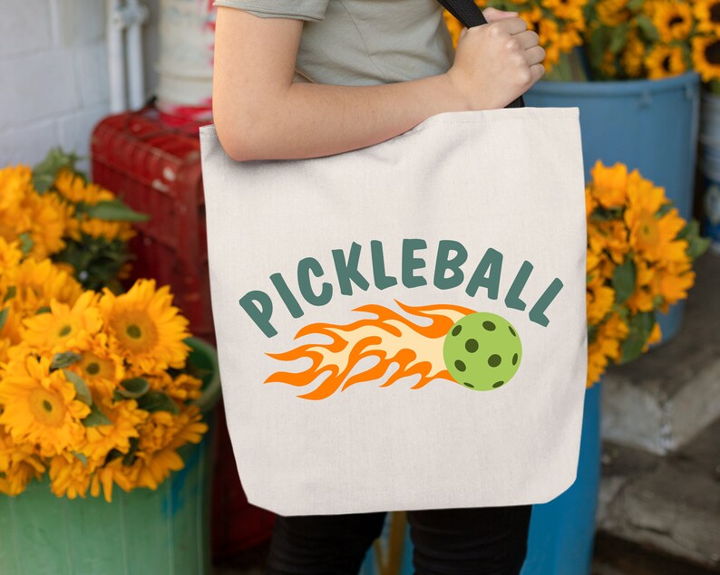 May include: A white canvas tote bag with a green and orange graphic of a pickleball on fire and the text "PICKLEBALL".