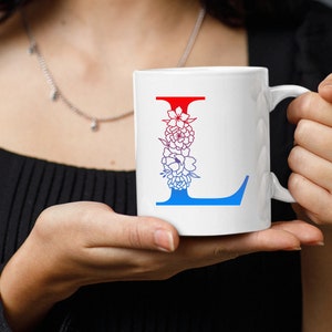 May include: White ceramic mug with a blue and red floral design featuring the letter L. The mug has a handle.