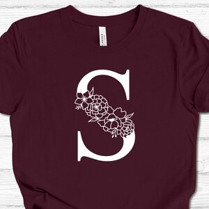 May include: A maroon t-shirt with a white letter "S" decorated with white floral designs.