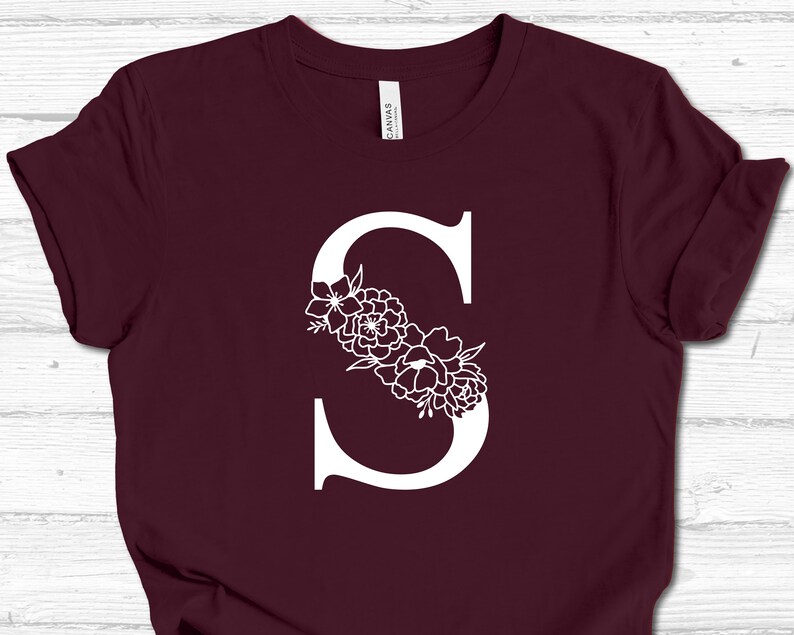 May include: A maroon t-shirt with a white letter "S" decorated with white floral designs.