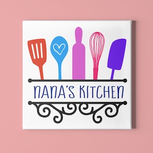 May include: A white canvas with a black sign that says "NANA'S KITCHEN".  The sign has a decorative scroll design and is hanging from a black rod.  There are five colorful kitchen utensils hanging from the rod: a red spatula, a blue spoon with a white heart, a pink rolling pin, a pink whisk, and a purple spatula.