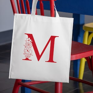 May include: White canvas tote bag with a red letter M and a floral design. The bag is hanging on a red chair.
