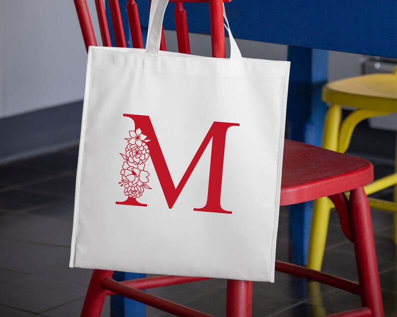 May include: White canvas tote bag with a red letter M and a floral design. The bag is hanging on a red chair.