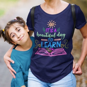 May include: A woman and a young girl wearing blue shirts. The woman's shirt has a graphic design that says "It's a beautiful day to learn" with a sun, book, and pencil.