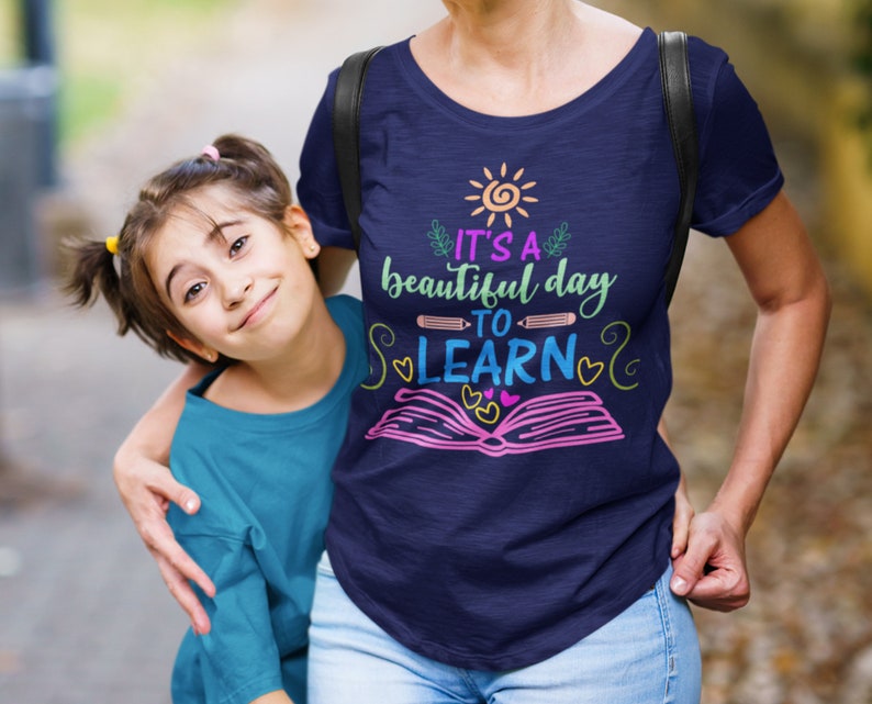 May include: A woman and a young girl wearing blue shirts. The woman's shirt has a graphic design that says "It's a beautiful day to learn" with a sun, book, and pencil.