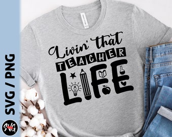 Op de afbeelding: Grijs T-shirt met zwarte tekst die "Livin' that teacher life" luidt. De tekst wordt omringd door verschillende schoolgerelateerde pictogrammen, waaronder een potlood, een gloeilamp, een boek en een appel.