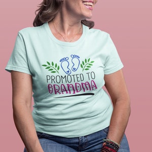 May include: A light blue t-shirt with a graphic design of two blue baby footprints with green leaves and the text "Promoted to Grandma" in pink and black.