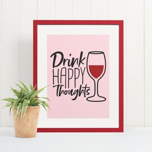 May include: A framed print with a pink background and black text that reads "Drink Happy Thoughts". A black outline of a wine glass with red wine is to the right of the text.