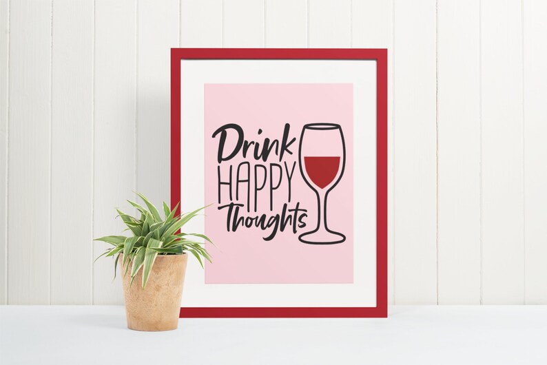 May include: A framed print with a pink background and black text that reads "Drink Happy Thoughts". A black outline of a wine glass with red wine is to the right of the text.