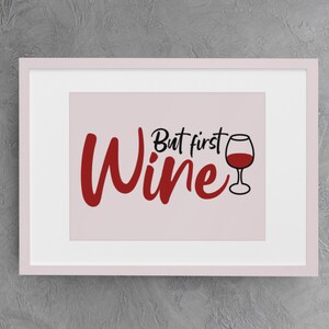 May include: A white framed print with a pink background and the text "But first, wine" in black and red lettering. A red wine glass is pictured to the right of the text.