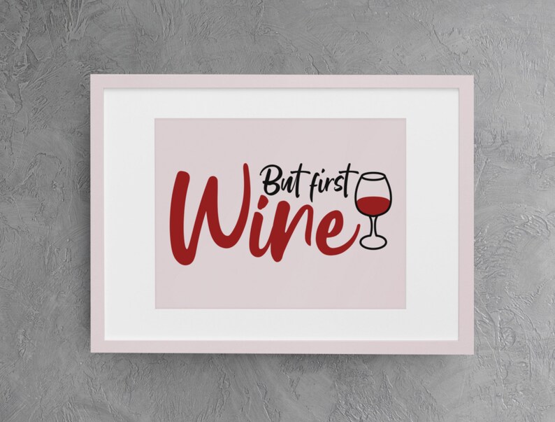 May include: A white framed print with a pink background and the text "But first, wine" in black and red lettering. A red wine glass is pictured to the right of the text.