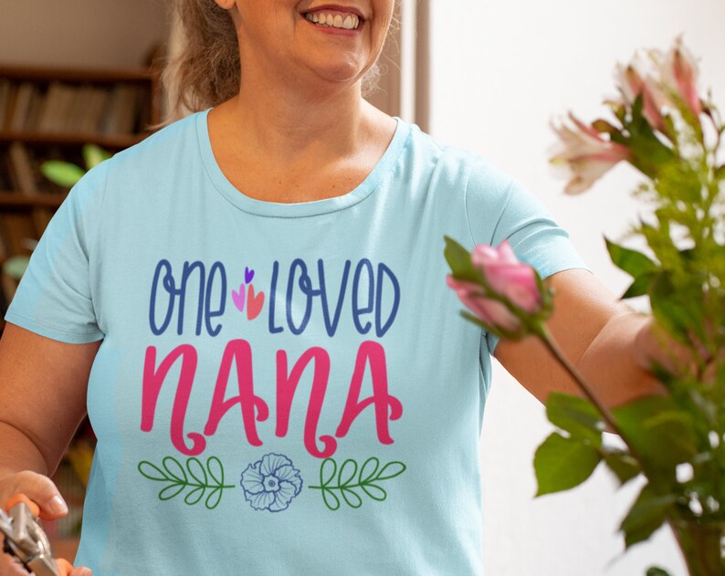May include: A light blue t-shirt with the text "one loved nana" in pink and blue lettering. The shirt has a floral design with green leaves and a blue flower.