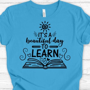 May include: A light blue t-shirt with a black graphic design that says "It's a beautiful day to learn". The graphic includes a sun, a book, pencils, and hearts.