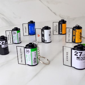 May include: A collection of film canister keychains with personalized messages. The canisters are black, green, and yellow, with white labels. Some labels read "Happy Anniversary!" and "Our First Trip."