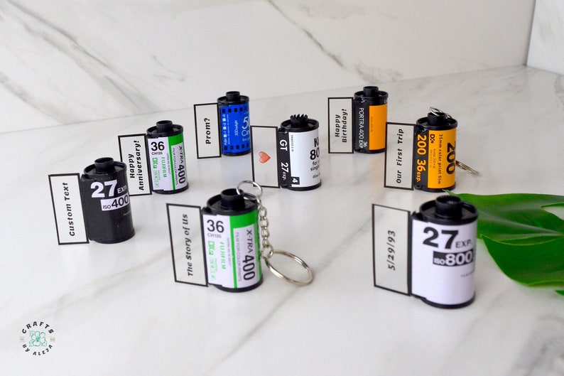 May include: A collection of film canister keychains with personalized messages. The canisters are black, green, and yellow, with white labels. Some labels read "Happy Anniversary!" and "Our First Trip."