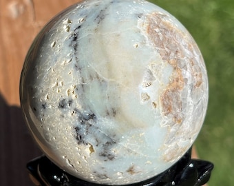 May include: A smooth, white and gray sphere with brown and black markings, resting on a black stand. The sphere is made of polished stone and has a natural, organic look.