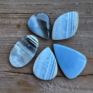 May include: Five polished gemstones in shades of blue, gray, and white are arranged on a wooden surface. The stones have various shapes, including teardrop and leaf-like forms. Some stones display banded patterns, while others are solid blue.