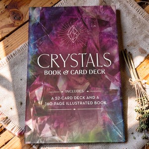 May include: A rectangular book and card deck set titled Crystals Book & Card Deck. The cover features a purple and green geometric crystal pattern with a white line-art crystal icon. It includes a 52-card deck and a 160-page illustrated book.