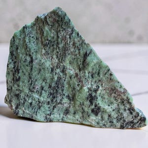 May include: A rough, triangular-shaped mineral specimen featuring a mottled texture with shades of light green, dark green, and black. The surface displays natural crystalline patterns and variations in color density across its jagged edges.