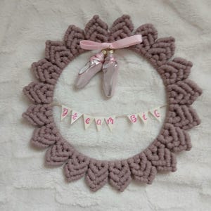 Puede incluir: Una corona de macramé hecha a mano en color malva apagado, con un diseño decorativo. La corona está adornada con una cinta rosa, dos zapatillas de ballet y una pancarta que dice "dream big". Una encantadora pieza de decoración.