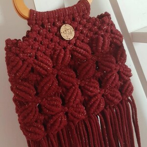 May include: A handmade burgundy macrame handbag with a wooden handle. The bag features a textured pattern and a fringe detail at the bottom. A small wooden button with text is attached to the front.