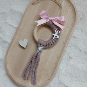 Puede incluir: Un adorno decorativo con temática de ballet. Presenta un anillo de madera envuelto en hilo malva, adornado con un dije de bailarina plateada y un lazo de satén rosa. Una borla cuelga debajo, y también hay un pequeño corazón de madera.