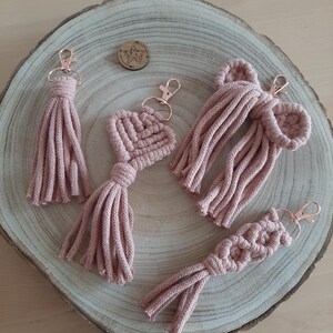 Può includere: Una collezione di portachiavi macramè fatti a mano in rosa tenue, con design a nappa, cuore e fiocco. I portachiavi hanno chiusure color oro rosa. Sono esposti su una superficie di legno.