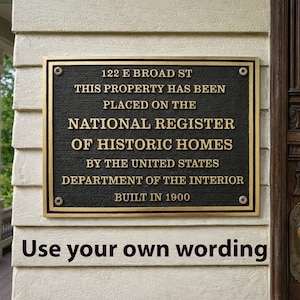 May include: A rectangular bronze plaque with raised gold lettering mounted on a cream-colored wall. The text reads: 122 E BROAD ST, THIS PROPERTY HAS BEEN PLACED ON THE NATIONAL REGISTER OF HISTORIC HOMES BY THE UNITED STATES DEPARTMENT OF THE INTERIOR, BUILT IN 1900.