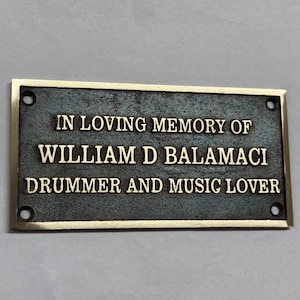 May include: A rectangular bronze plaque with a gold border. The text "IN LOVING MEMORY OF WILLIAM D BALAMACI DRUMMER AND MUSIC LOVER" is engraved in a serif font. Four holes are drilled in each corner.