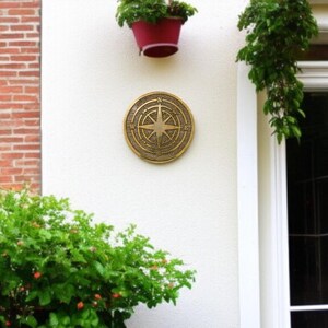 May include: A round, bronze-colored compass rose plaque mounted on a white wall. The plaque features directional markings and a central star design. The wall is adjacent to a brick wall and a white-framed window.
