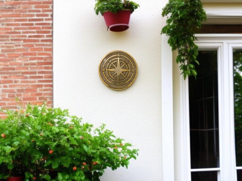 May include: A round, bronze-colored compass rose plaque mounted on a white wall. The plaque features directional markings and a central star design. The wall is adjacent to a brick wall and a white-framed window.