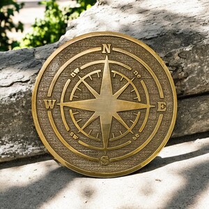 May include: A round, bronze-colored compass rose with directional indicators. The compass features a central star design and is set against a textured background. The letters N, E, S, and W mark the cardinal directions.