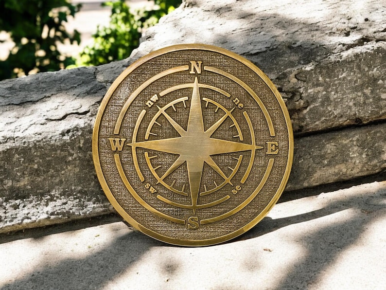 May include: A round, bronze-colored compass rose with directional indicators. The compass features a central star design and is set against a textured background. The letters N, E, S, and W mark the cardinal directions.